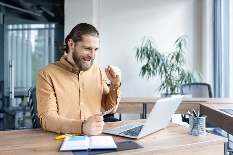 A man, excited, holds a credit card while sitting in front of a laptop at a modern office desk. Celebrating a successful online transaction.